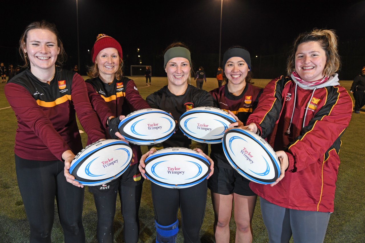 A group of female rugby players in burgundy team jackets holding out white rugby balls with the Taylor Wimpey logo on them.