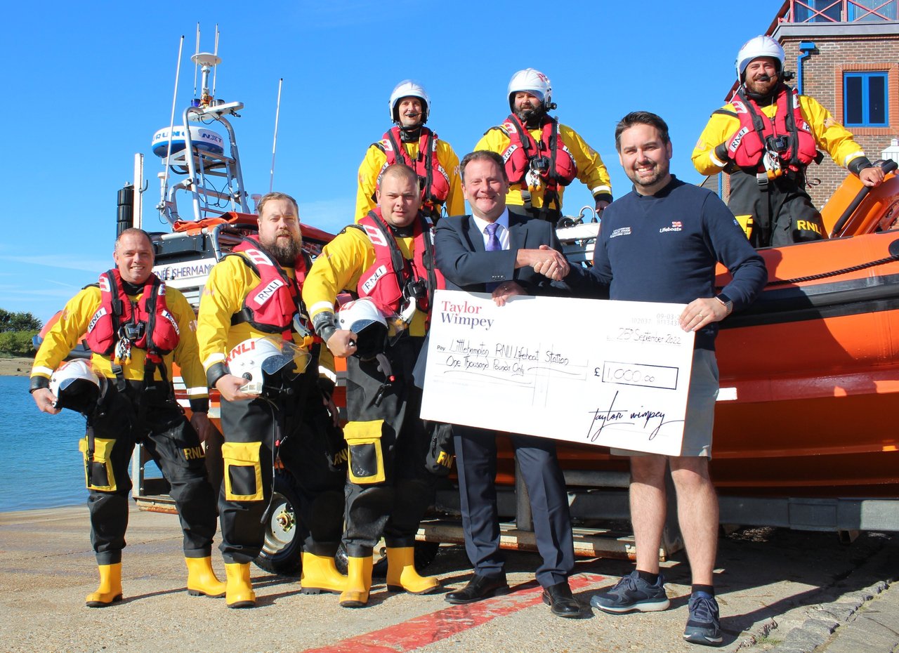 Two people holding a large cheque from Taylor Wimpey noting a donation to the Royal National Lifeboat Institution with a group of lifeguards in shot, next to water in front of clear blue sky.