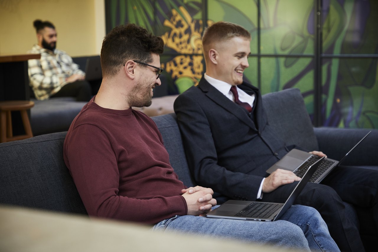 Two people, sat on a sofa with laptops on their laps. One is wearing a red jumper and blue jeans, and one is wearing a navy suit with a white shirt and dark red tie.