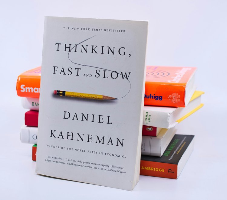 Daniel Kahneman's book, 'Thinking Fast and Slow', leaning against a pile of other books