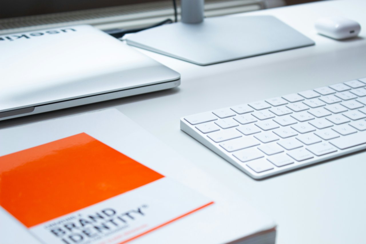 An orange book, entitled Brand Identity, sits on a white desk next to a white computer keyboard