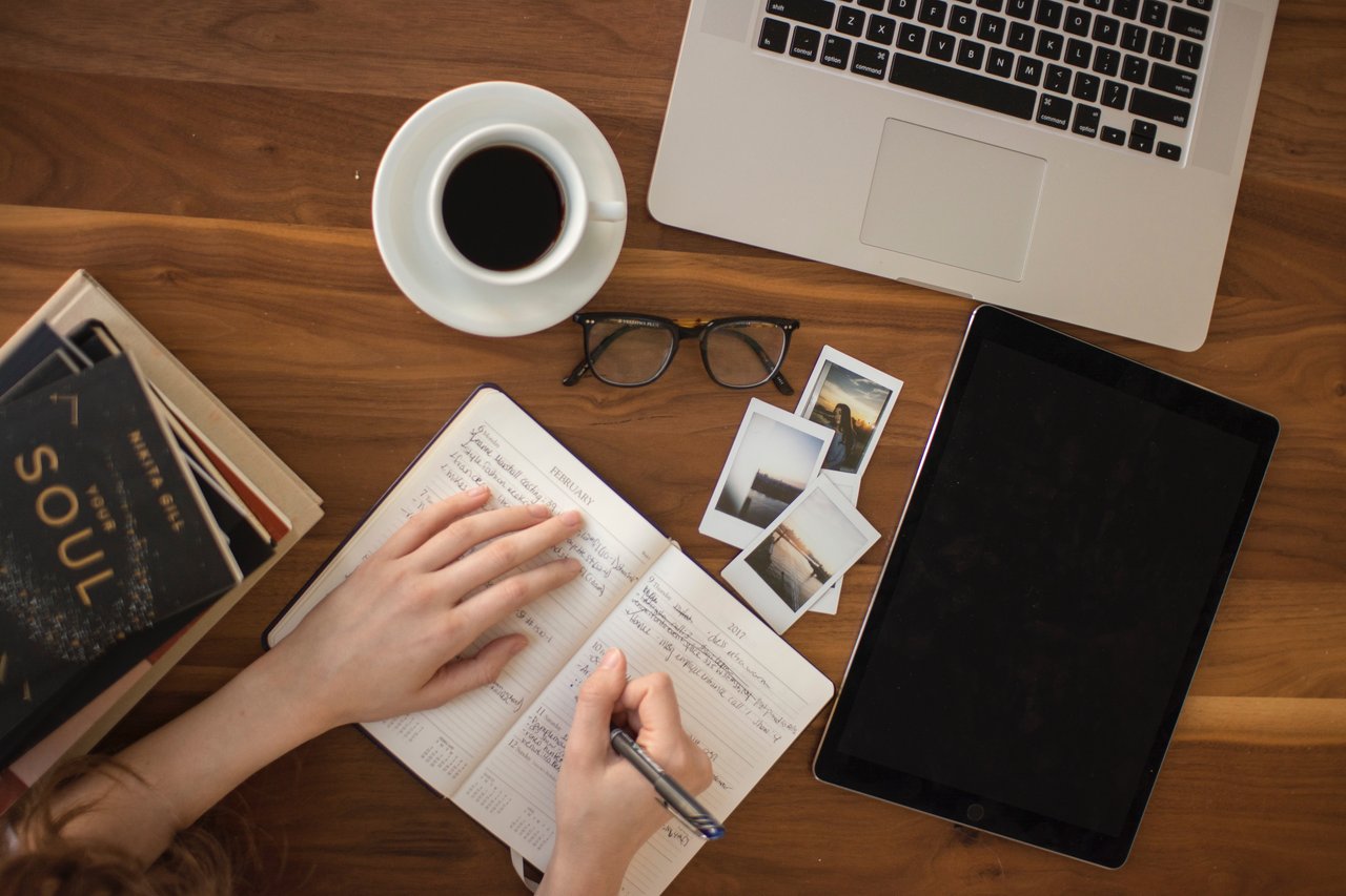 Someone writing on a notepad, with a cup of coffee, a laptop, some books and some photos around them.